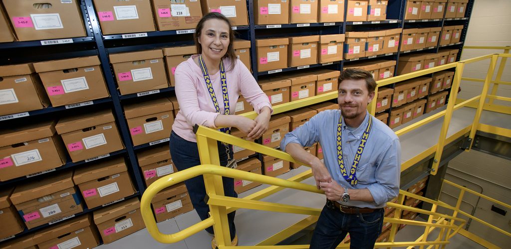 A male archivist stands on yellow stairs, while a female archivist stands near the stair railing. Behind them are a wall of boxes from the Bentley's collections.