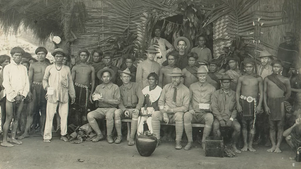 Two American colonial officials sitting with a group of Ifugao men in the Philppines.