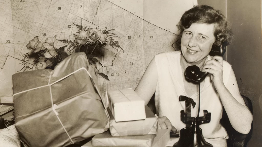 Josephine Gomon talking on a telephone with a pile of packages in front of her.