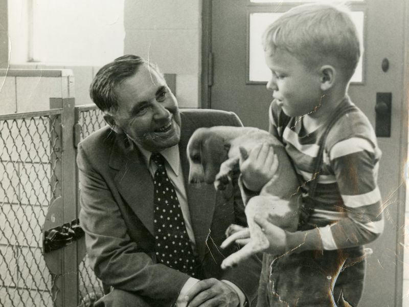 A man in a suit and tie crouching to smile at a boy in a striped shirt holding a puppy.