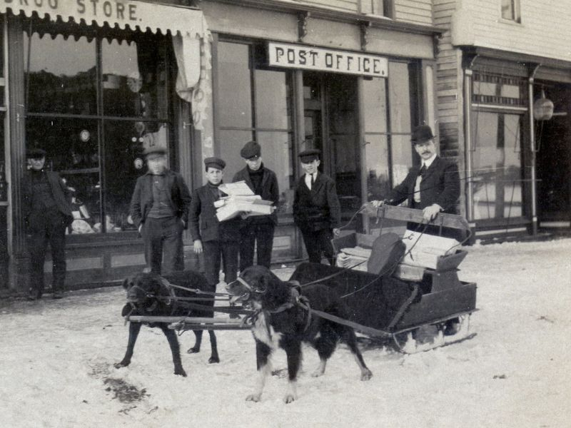 Two dogs pulling a mail sled, posed with four men in front of a Post Office in the snow.