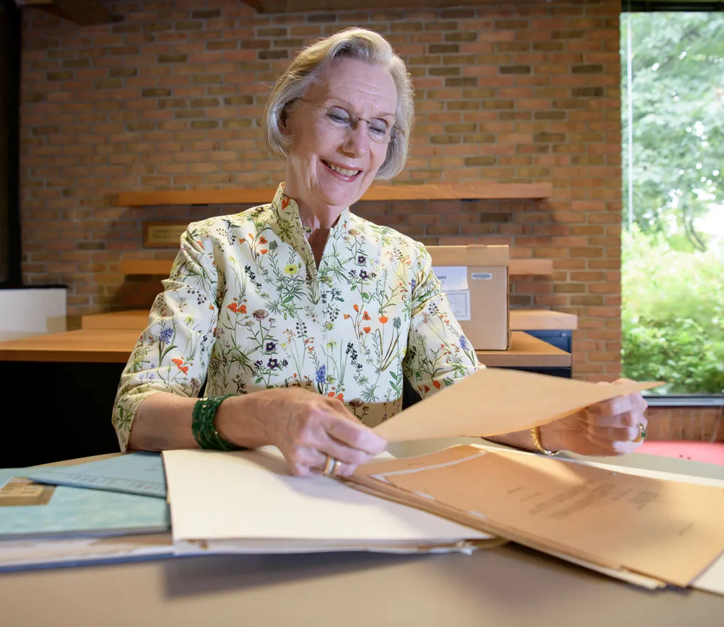 A woman with light-colored hair wearing a flowered shirt sits at a desk holding a folder of historical materials.