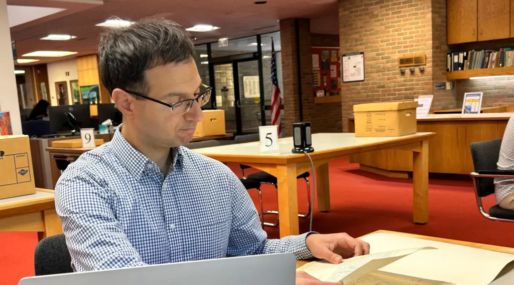 A visiting fellow to the Bentley Historical Library looking through historical papers at a table in the reading room, wearing a blue and white checkered shirt and glasses, with a laptop open in front of him. Several tables holding boxes from the archives are shown in the background behind him, ready for other researchers to arrive. The front desk, the red carpet of the reading room, and the library's brick walls are also visible.