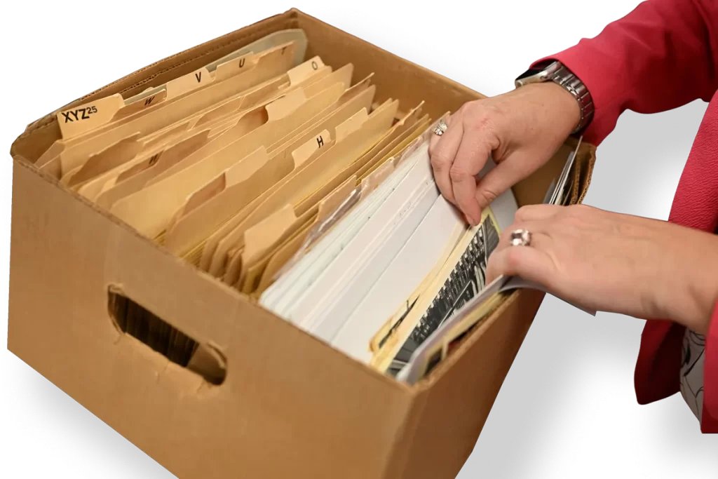 A researcher looking through folders of historical photos at the Bentley Historical Library. Only the researcher's hands and arms are visible, wearing a bright pink jacket, a watch, and two rings, reaching out to flip through folders in an open box.