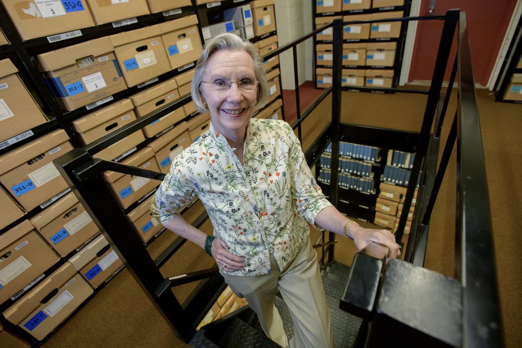 A woman wearing tan pants and a flower-patterned shirt stands on a set of black stairs surrounded by brown archival boxes on shelves.
