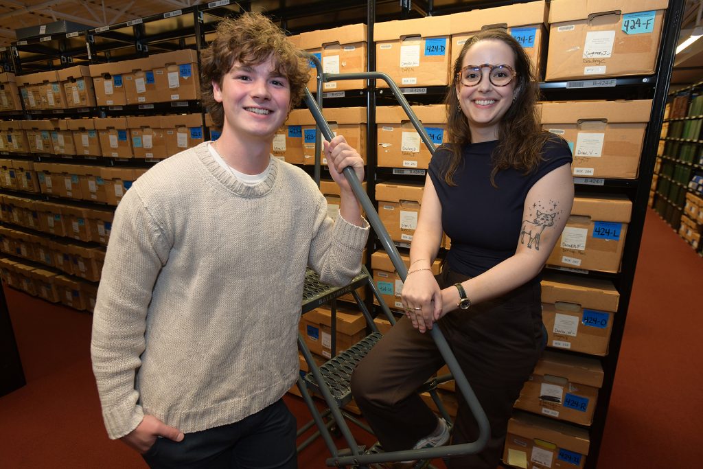 A man and woman in the stacks of the Bentley Historical Library, surrounded by boxes on shelves. The man is on the left wearing a light-colored sweater, the woman is on the right wearing a dark top.