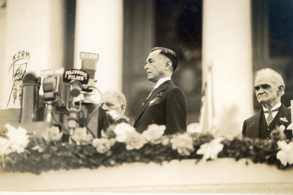 A man with dark hair wearing a dark suit jacket stands in front of a cluster of microphones.